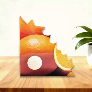 Cardboard box with a bright orange slice graphic and a circular window, sitting on a wooden table beside a potted plant.